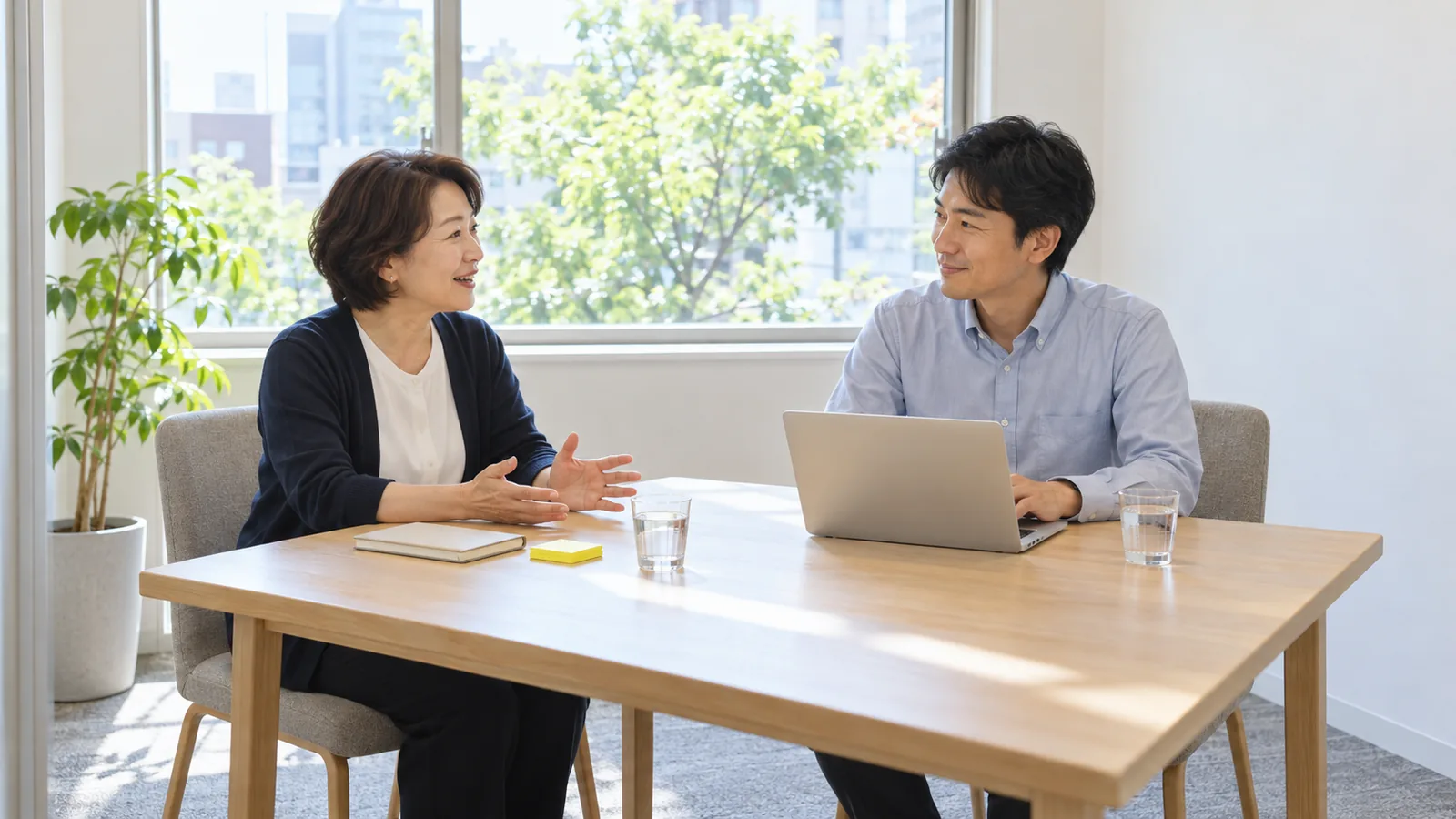 明るい会議室で、お店を経営する女性が自分の仕事を話し、向かいの男性がノートパソコンで書き留めている